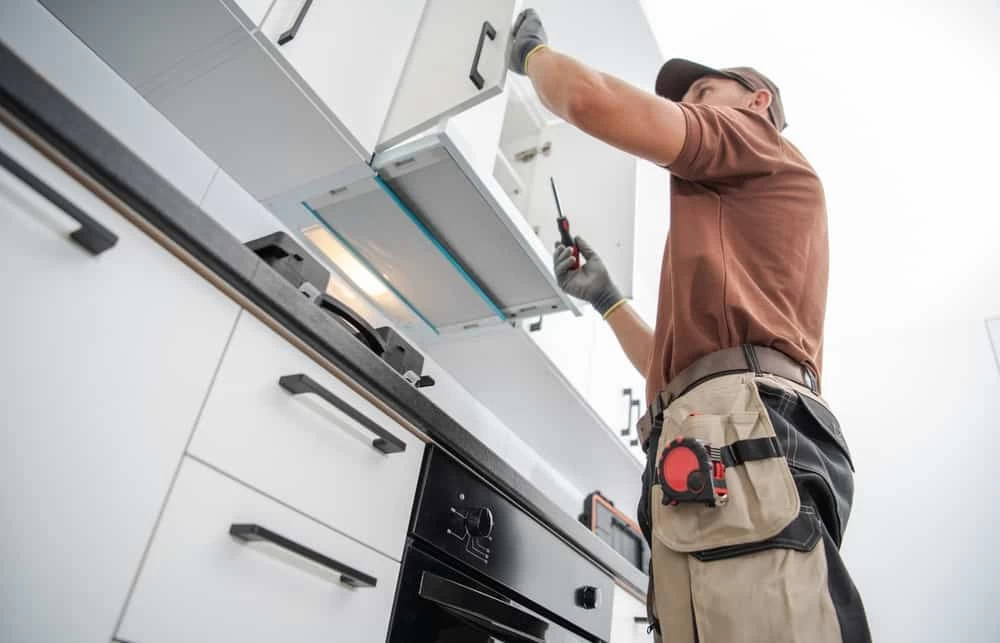 A maintenance worker in a brown shirt and protective gloves repairs kitchen cabinets with a screwdriver, as part of an ongoing kitchen renovation. Tools are attached to his utility belt.
