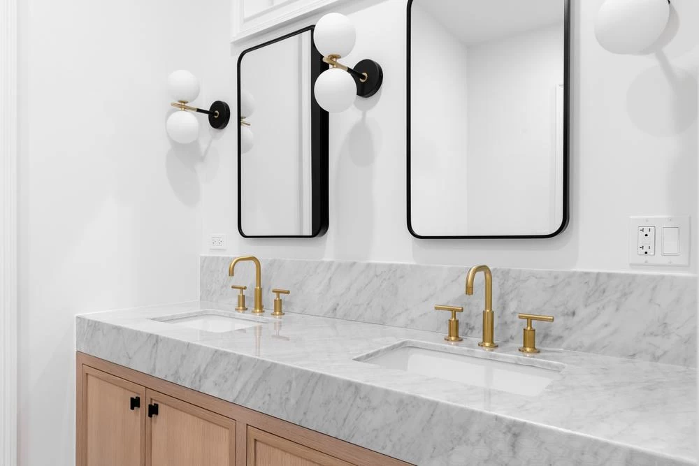 A modern bathroom with a double sink vanity, gold hardware, marble countertop, rectangular mirrors, and wall-mounted globe light fixtures showcases the finesse often seen in high-end home renovations.