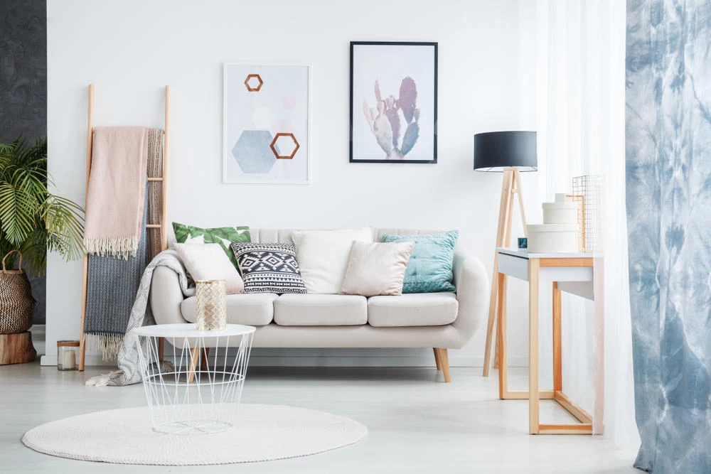 A cozy living room, perfect for a home remodeler, features a white sofa with colorful cushions, minimalist wall art, a ladder with blankets, a round coffee table, and a floor lamp.