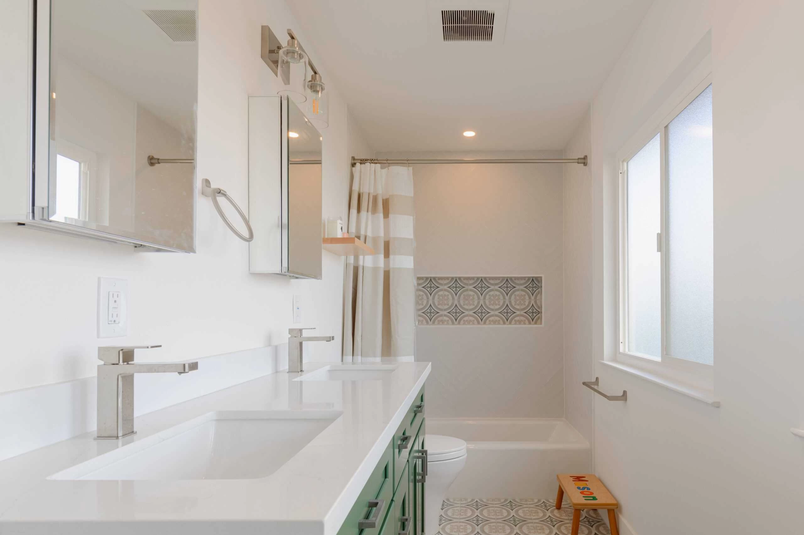 A modern bathroom with dual sinks, green cabinets, a bathtub with a patterned tile accent.