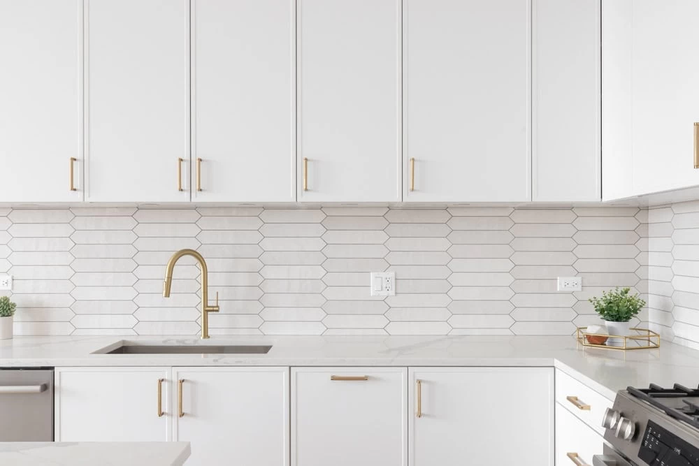 Modern kitchen with white cabinets, brass hardware, white geometric tile backsplash, stainless steel sink with brass faucet, and small plants.