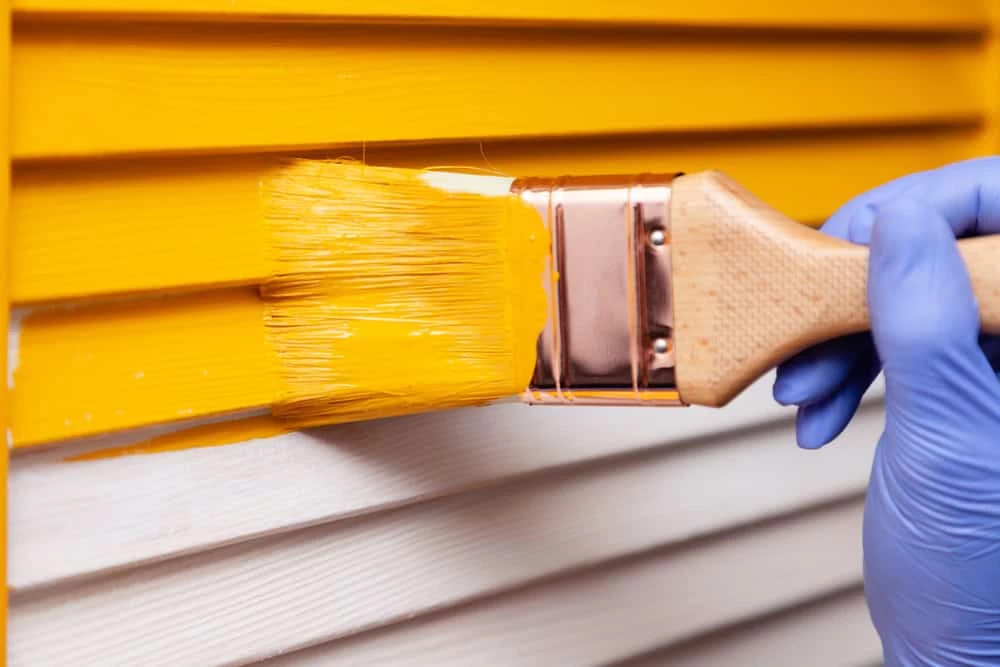 A person in blue gloves is painting a yellow siding.